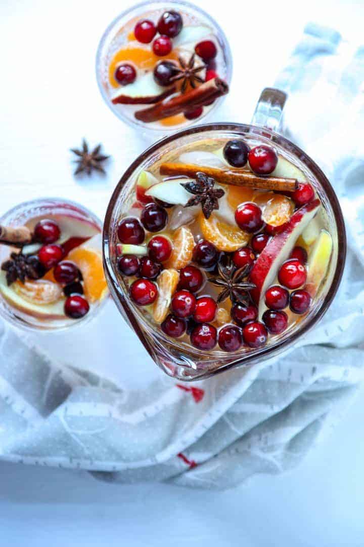 Top down view of two glasses and a carafe of winter white sangria filled with cinnamon sticks, apple slices, orange slices, and cranberries