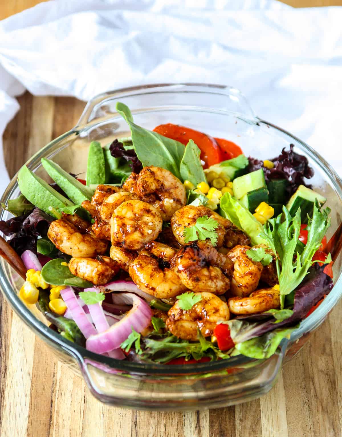 Salad topped with grilled shrimp in a glass bowl on a wooden board.