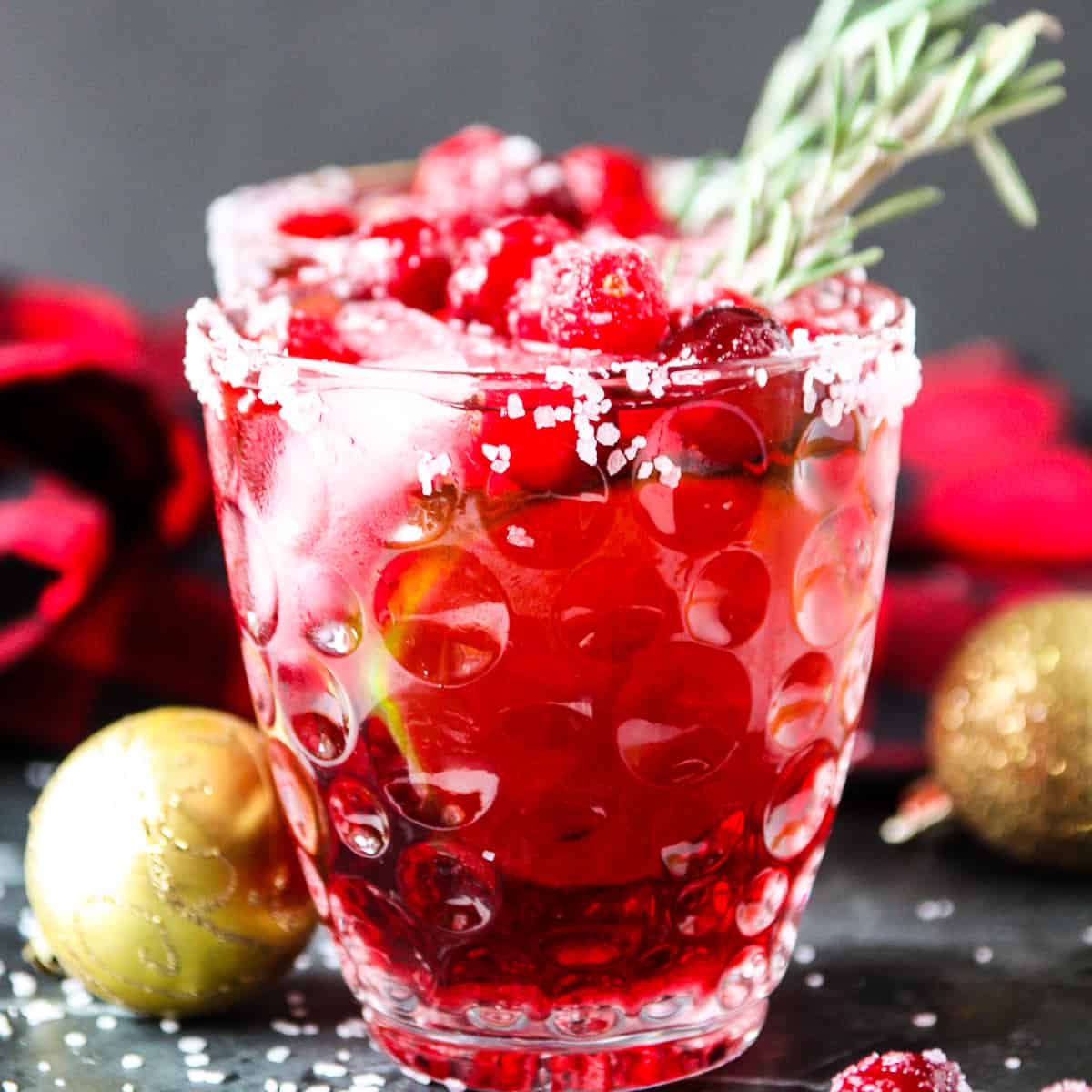 A cranberry mistletoe margarita drink in  a clear glass with a sprig of rosemary.
