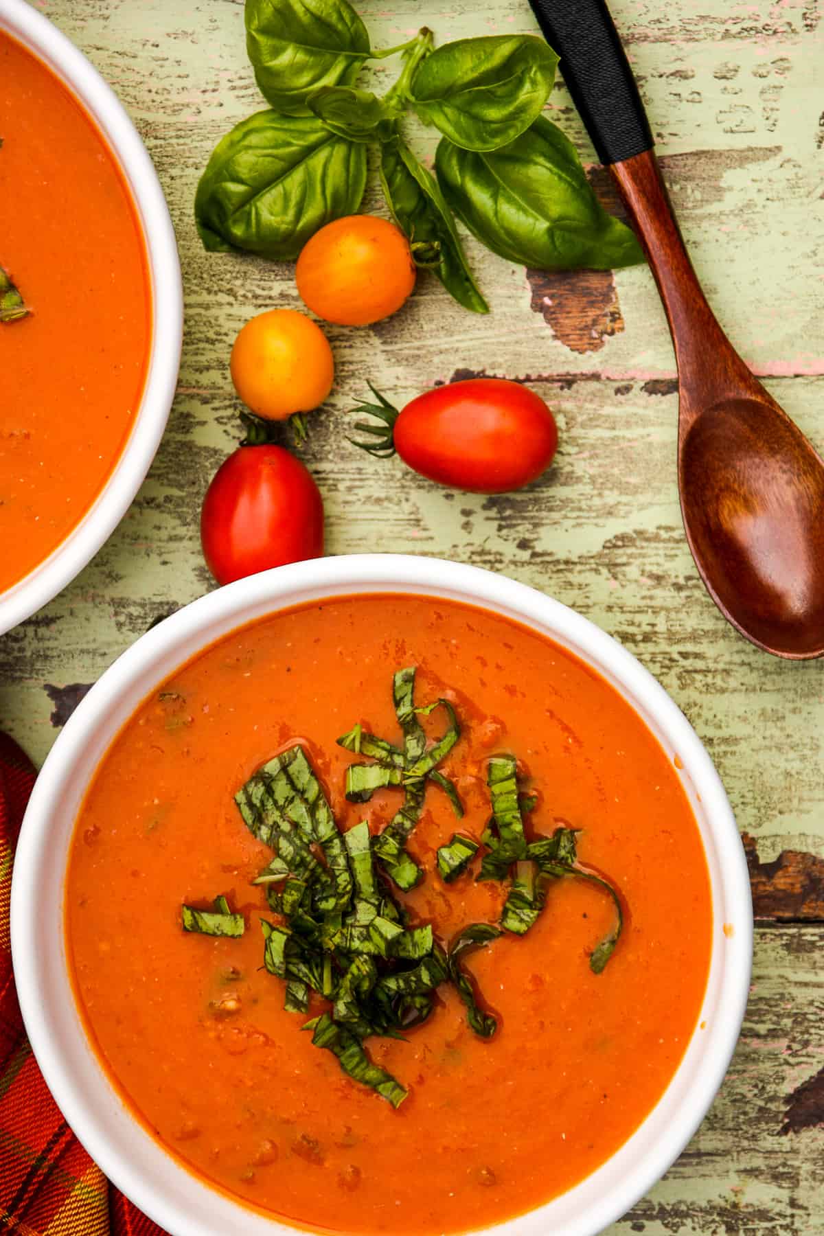 Two bowls of roasted tomato basil soup and two tomatoes on a weathered table.