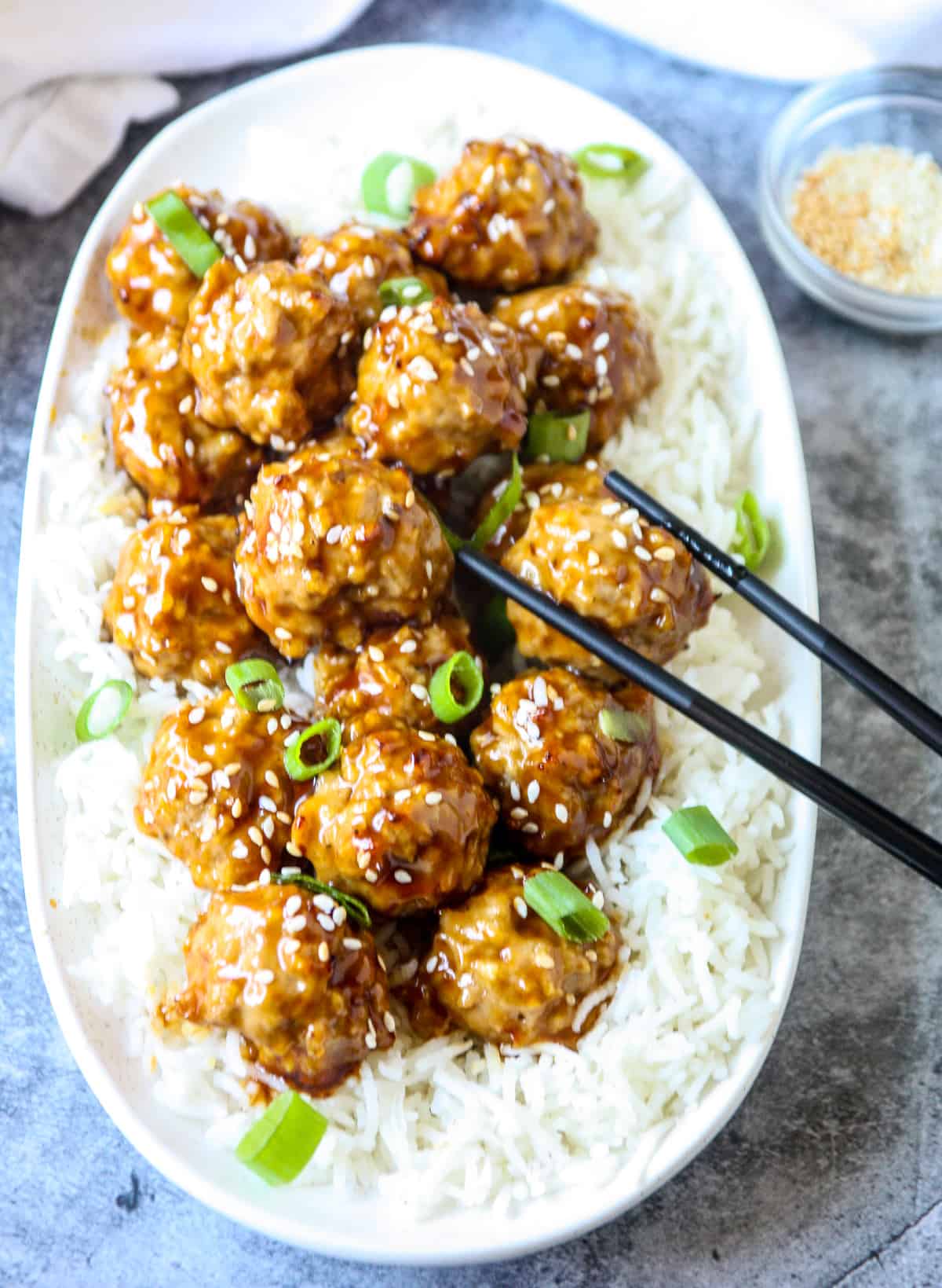Baked and glazed Asian chicken meatballs on top of rice on a white platter with black chopsticks.