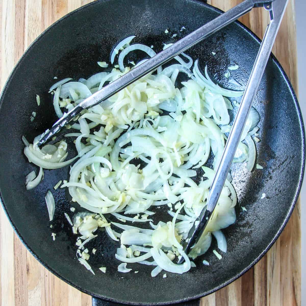 Onions and garlic being stir fried in a cast iron wok with metal tongs.
