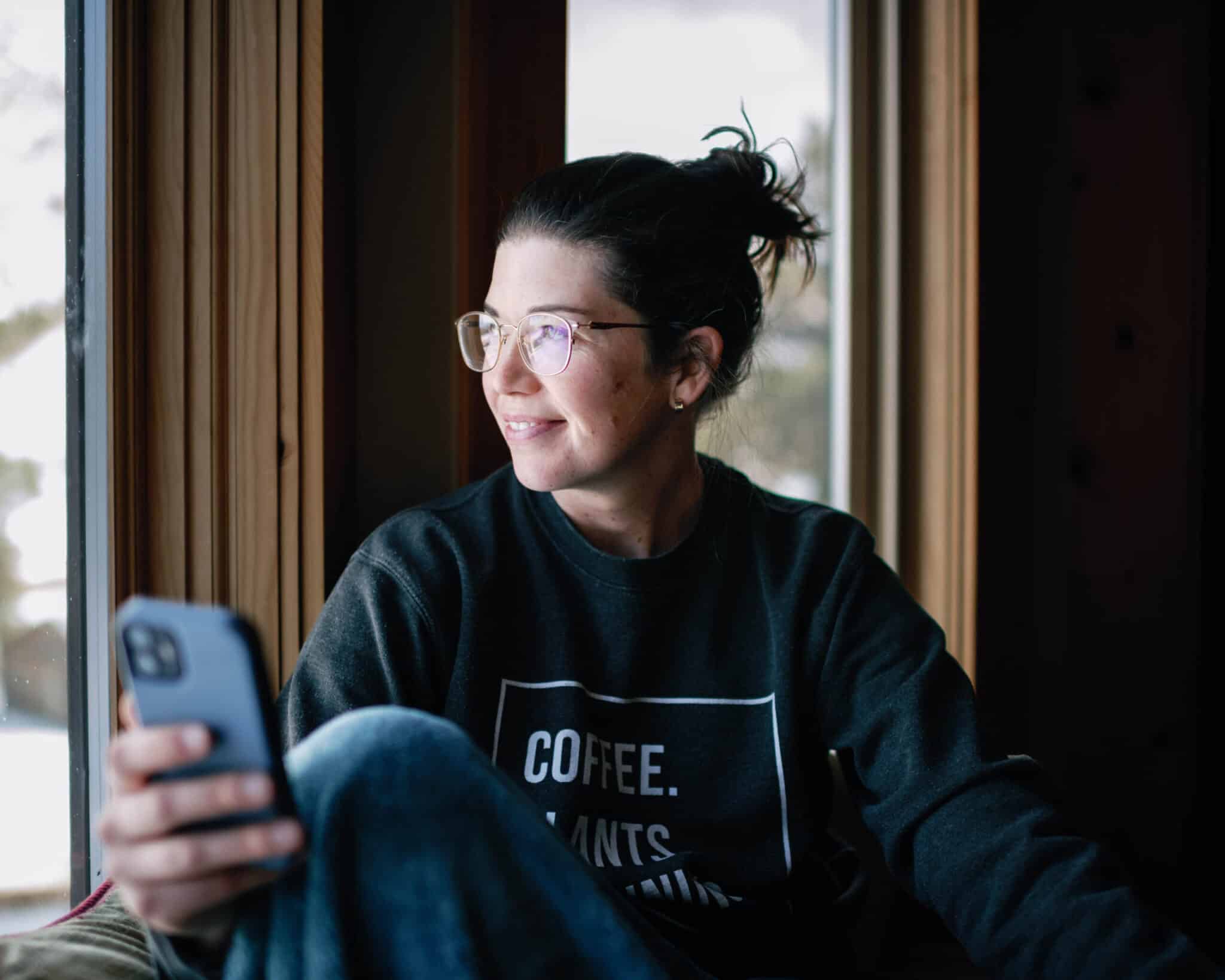 image of woman in comfy clothes looking out a window with phone in hand