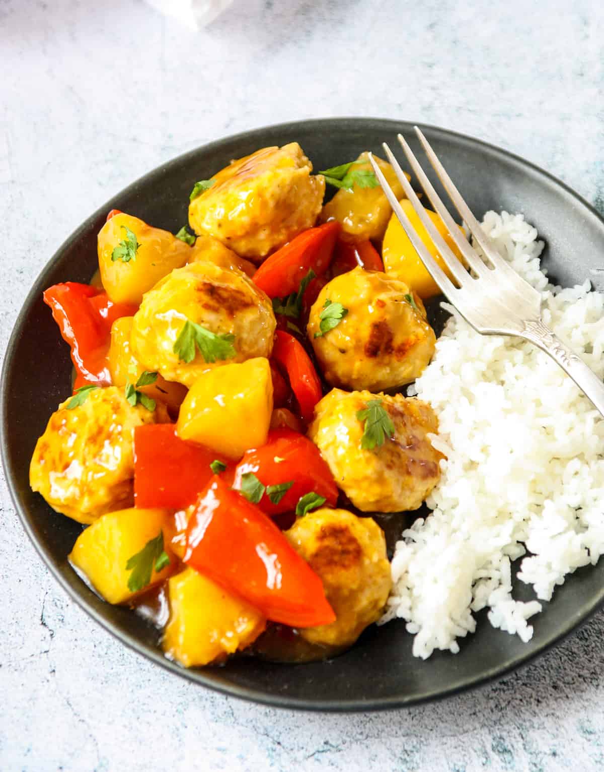 Meatballs with pineapple and red pepper on a black plate with rice and a silver fork.
