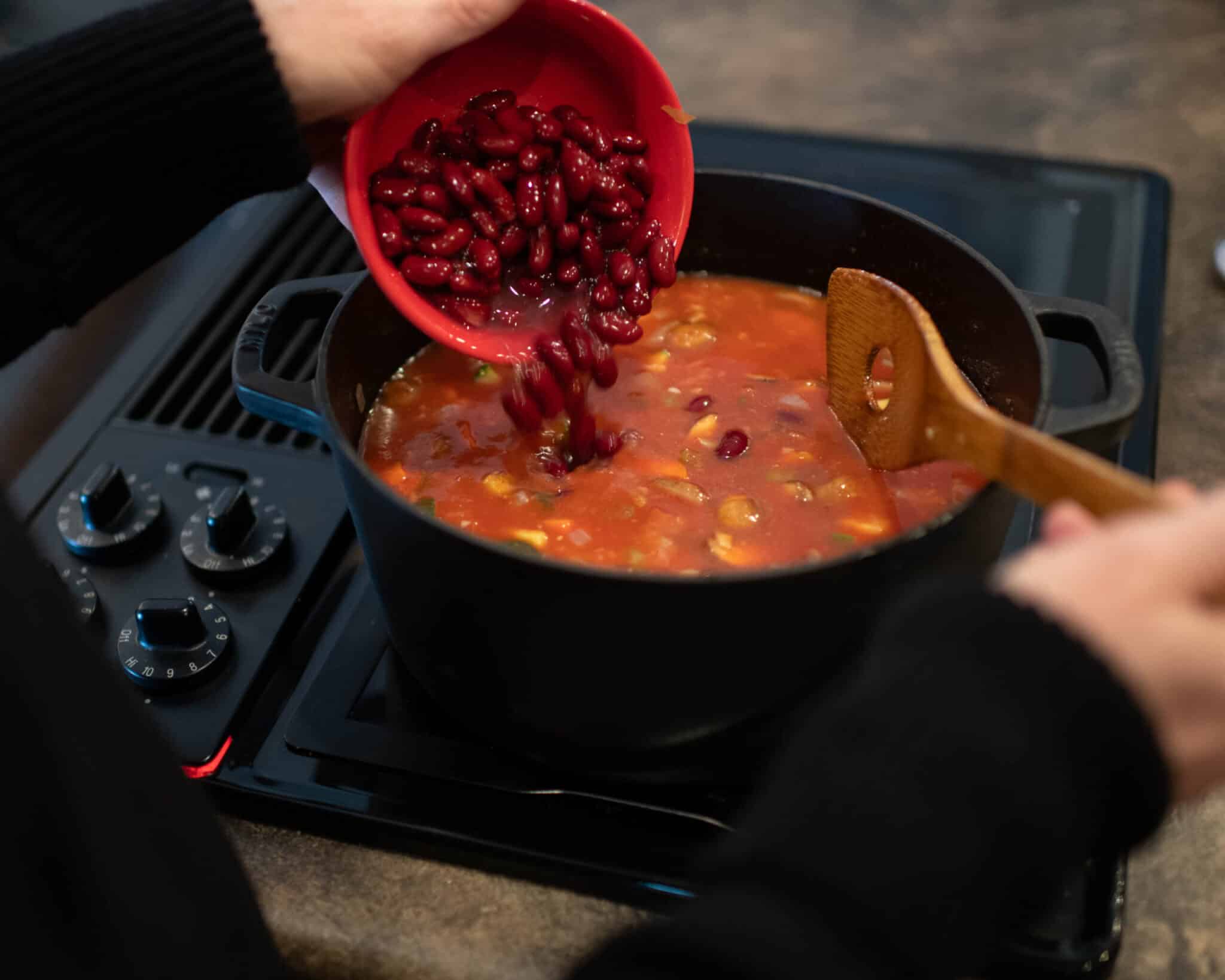 pouring kidney beans from a red bowl into a black pot of red stew